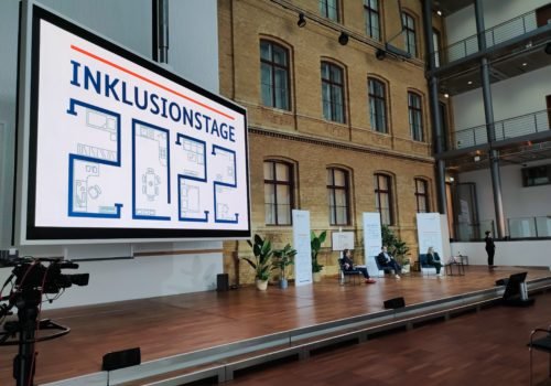 Foto des Podiums bei den Inklusionstagen. Links ein großer Bildschirm mit dem Schriftzug "Inklusionstage 2022"