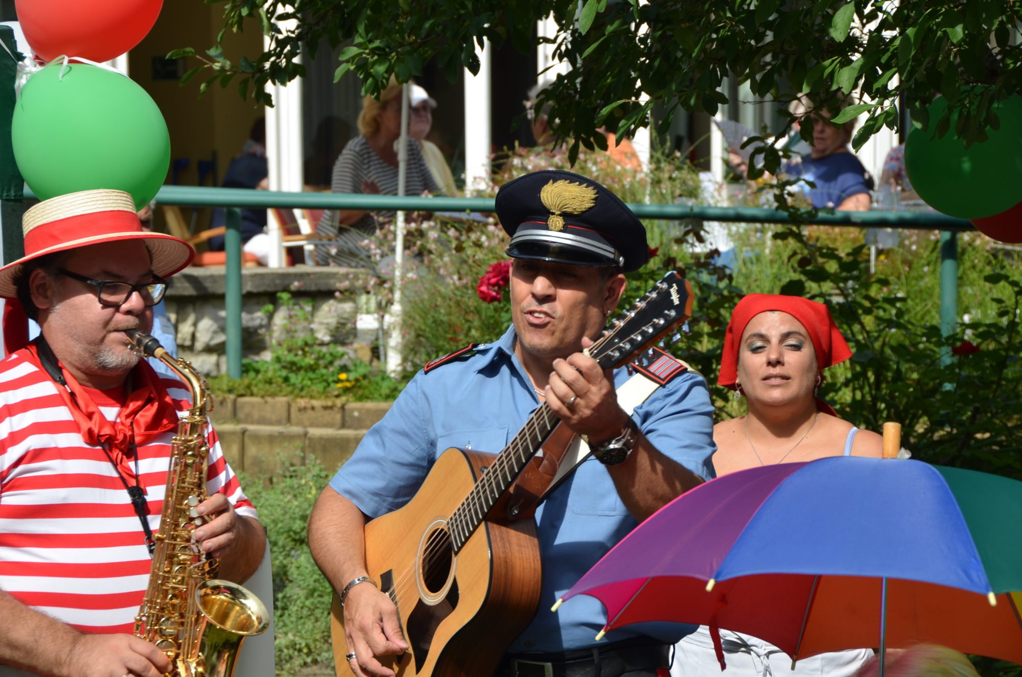 Drei Musiker - zwei Männer und eine Frau - spielen auf dem italiensichen Sommerfest der Villa Donnersmarck Musik. Ein Mann spielt Saxofon, ein weiterer Gitarre, die Frau singt.