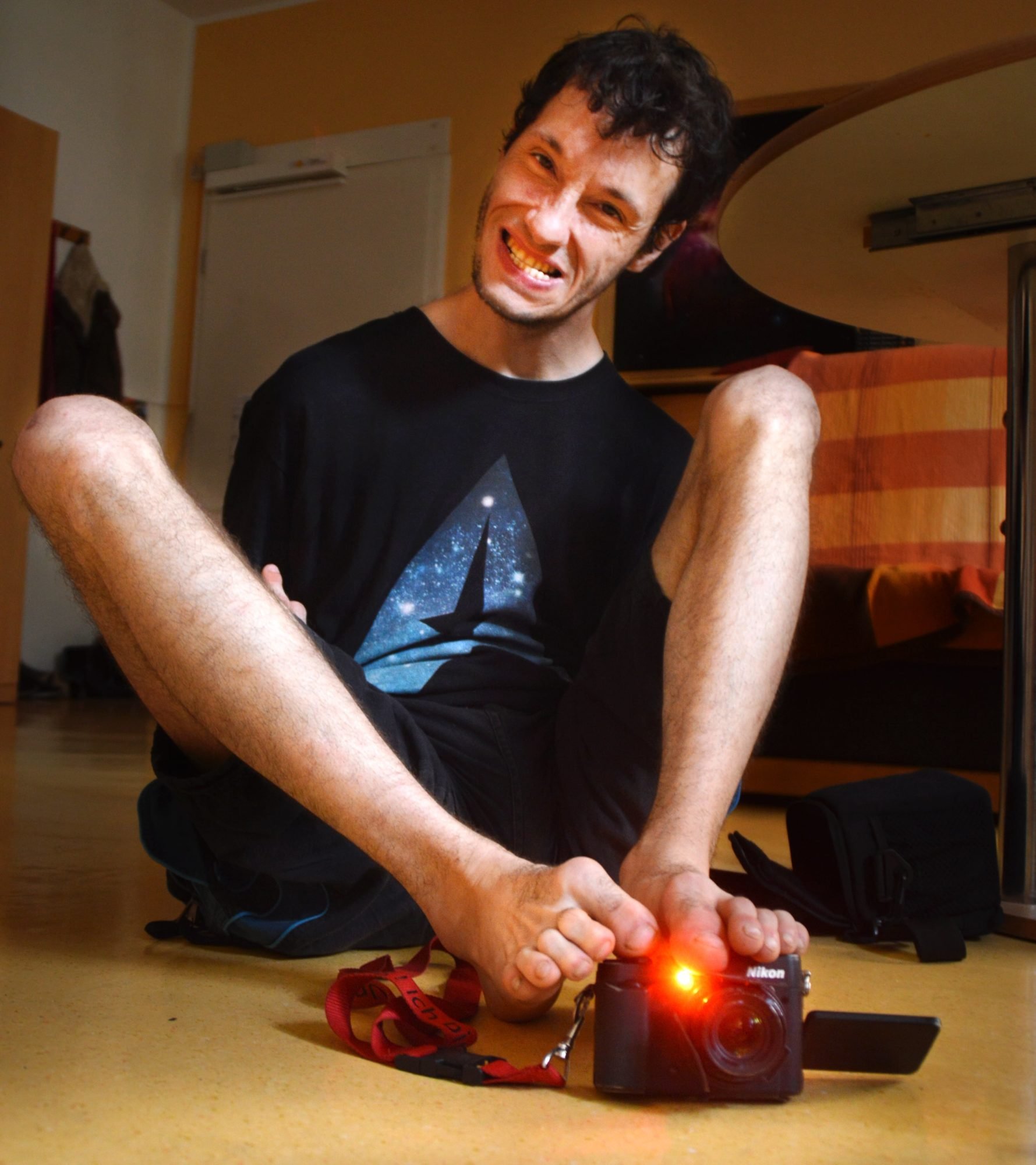 Fotograf Sven Kocar, lachend auf dem Fußboden seiner Wohnung sitzend, in seinen Füßen eine Digitalkamera, deren rotes aufnahmelämpchen leuchtet