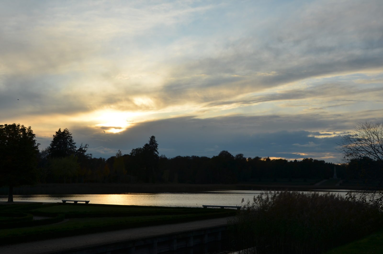 Ein Sonnenuntergang bei wolkenbedecktem Himmel, direkt am Wasser. Über den See hinweg sind im Hintergrund sind ein Wald und ein Obelisk.