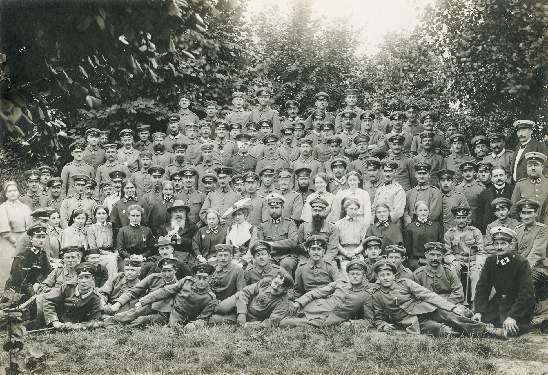 Eine Menschenmenge ist zu sehen. Die weitaus überwiegende Anzahl besteht aus Soldaten; teilweise sind Krücken zu sehen. In der Bildmitte sind jedoch gut situierte Personen zu erkennen - darunter Chefarzt Dr. Berg und Guido von Donnersmarck.