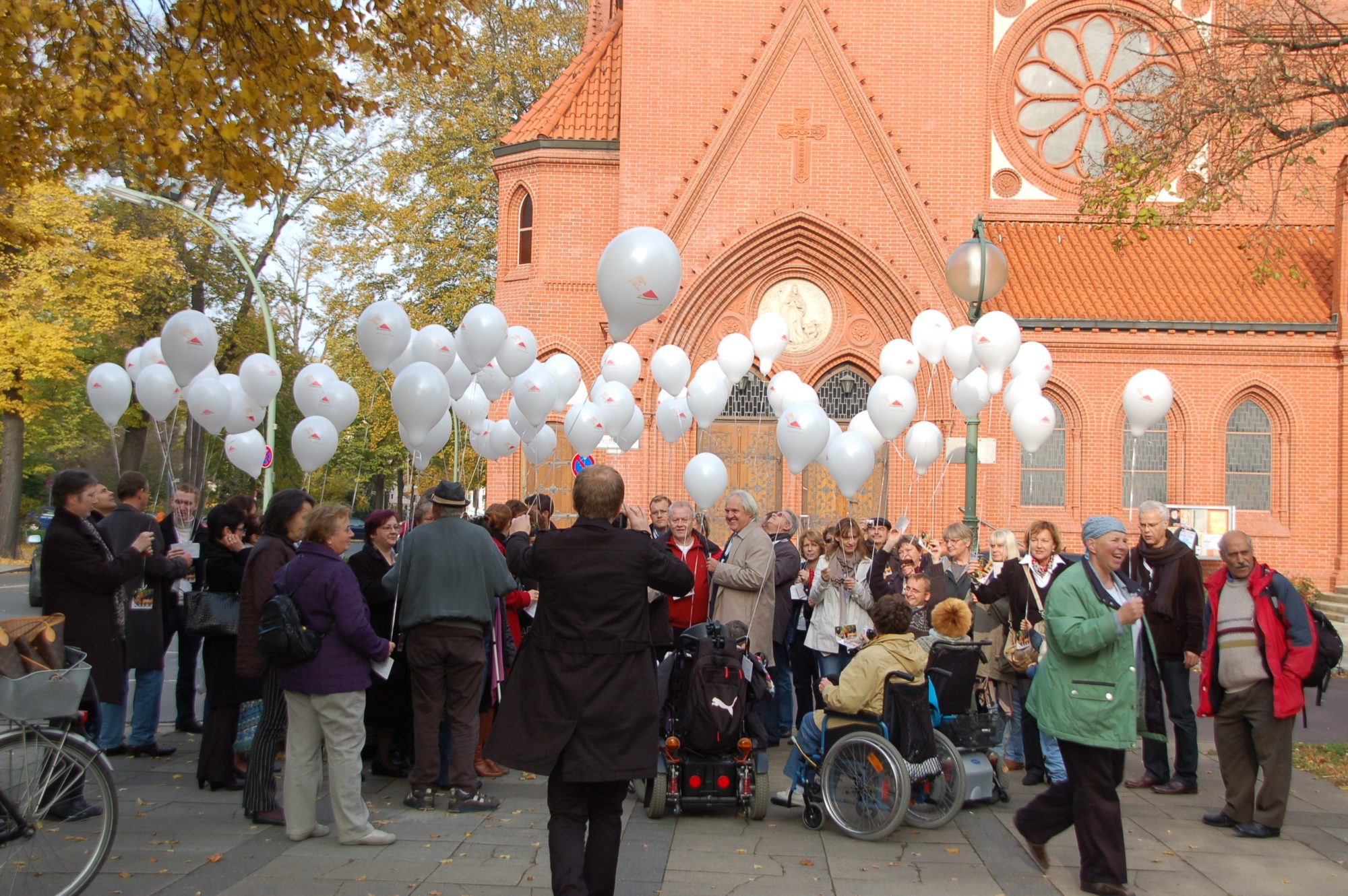 Eine große Menschentraube - zu Fuß und im Rollstuhl. Alle halten weiße Luftballons in den Händen.
