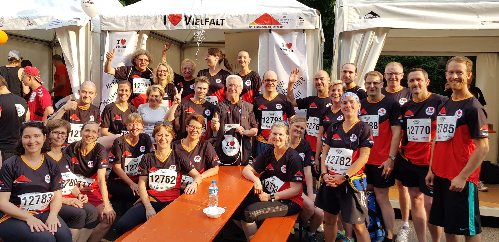 Mehrere Mitarbeiterinnen und Mitarbeiter im Laufdress stehen vor dem Zelt der Stiftung und winken in die Kamera.
