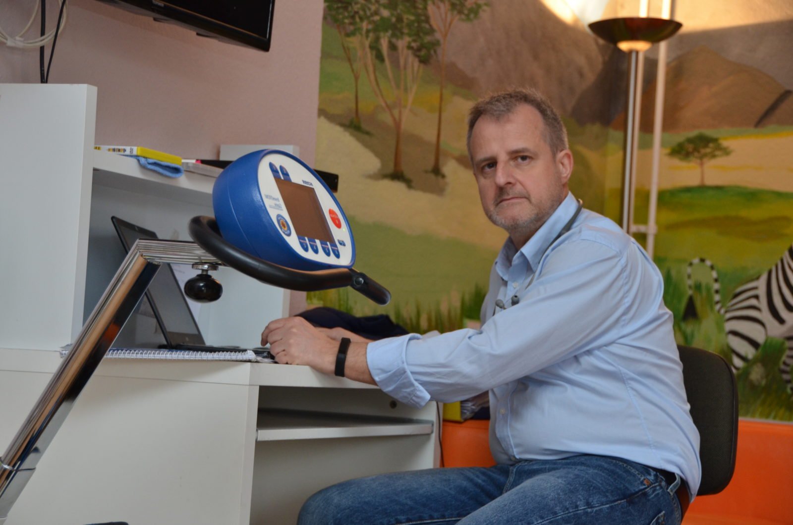 Michael Ertel sitzt am Schreibtisch, vor ihm ein Laptop und Notizblock, er hat ein Stethoskop um den Hals, im Vordergrund erkennt man ein Ergometer, im Hintergrund sieht man eine bemalte Wand mit einer Savannenszene und einem Zebra.