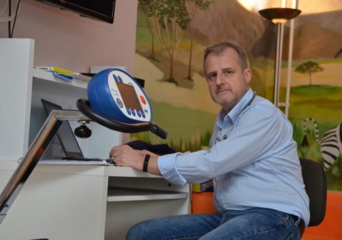 Michael Ertel sitzt am Schreibtisch, vor ihm ein Laptop und Notizblock, er hat ein Stethoskop um den Hals, im Vordergrund erkennt man ein Ergometer, im Hintergrund sieht man eine bemalte Wand mit einer Savannenszene und einem Zebra.
