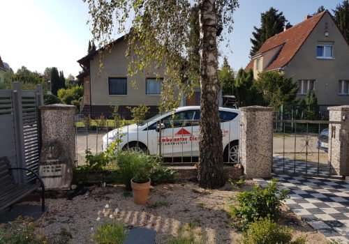 Ein Auto des Ambulanten Dienstes, mit Logos beklebt, steht in einem Wohngebiet am Straßenrand. Das Bild ist aus einem Vorgarten heraus fotografiert. Das Auto steht hinter einem Zaun, davor ein Baum und mehrere Töpfe mit Pflanzen.