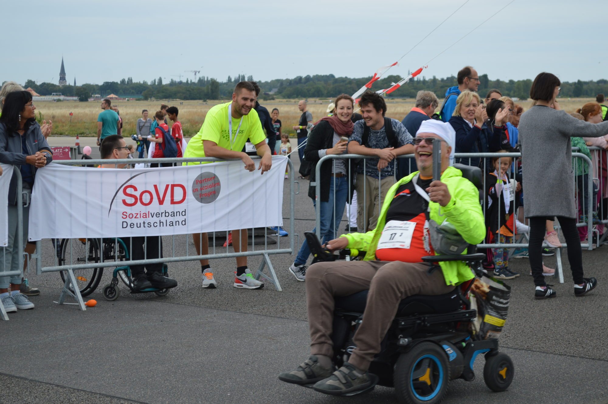 Rollifahrer aus dem Team FDST rollt mit seinem E-Rollstuhl an Zuschauern vorbei und steckt alle mit seinem Lächeln an.