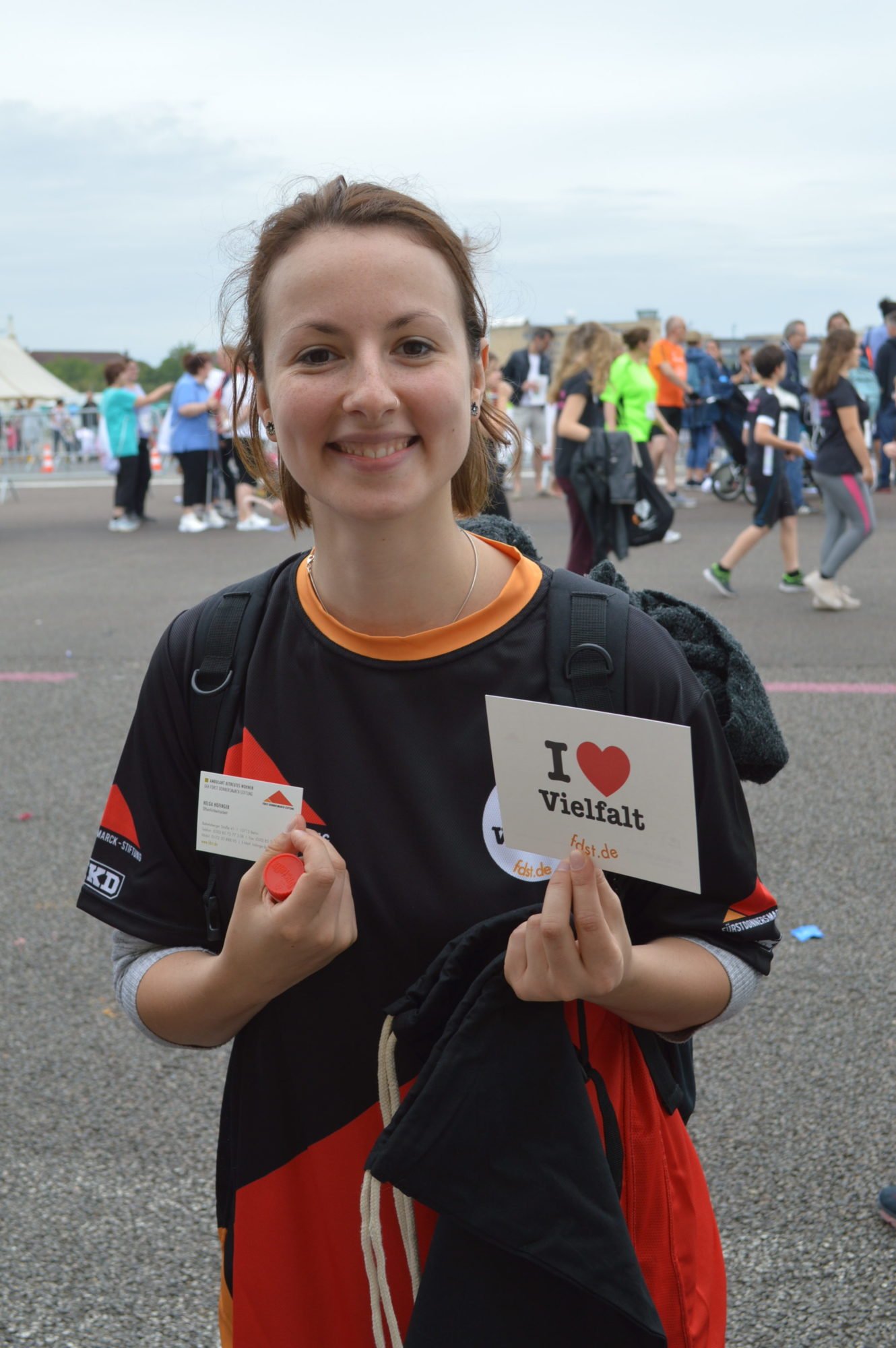 Eine junge Frau hält eine I love VIelfalt Karte nach oben, trägt eines unserer Team-Shirts und lächelt.