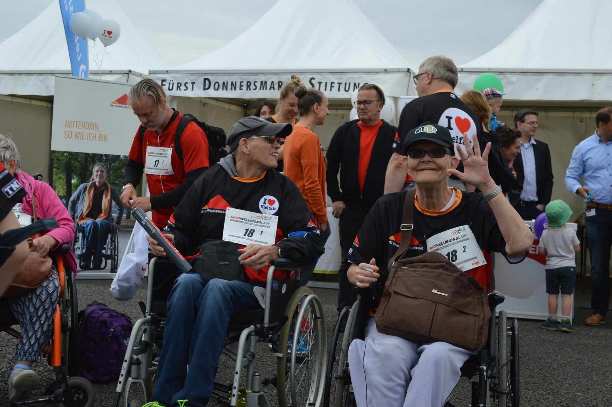 Rollstuhlfahrer und fahrerin vor dem Zelt der FDST, die Rollifahrerin winkt Richtung Kamera.