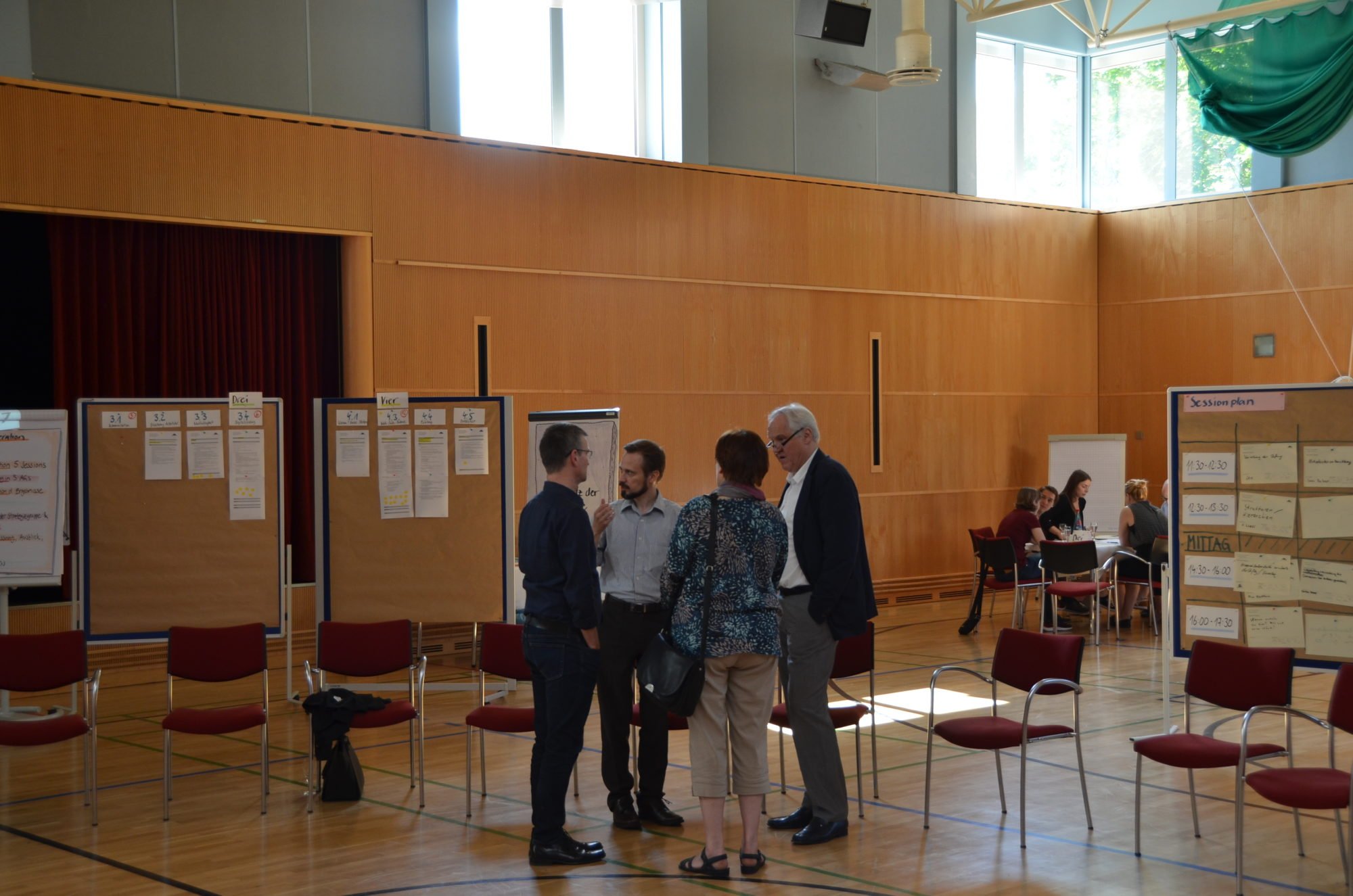 Die Next Generation-Tagung in der Sporthalle des Seehotel Rheinsberg. Wolfgang Schrödter spricht mit dem Führungskreis. Im Hintergrund sieht man Ergebnisse an Whiteboards und Kleingruppen im Austausch.
