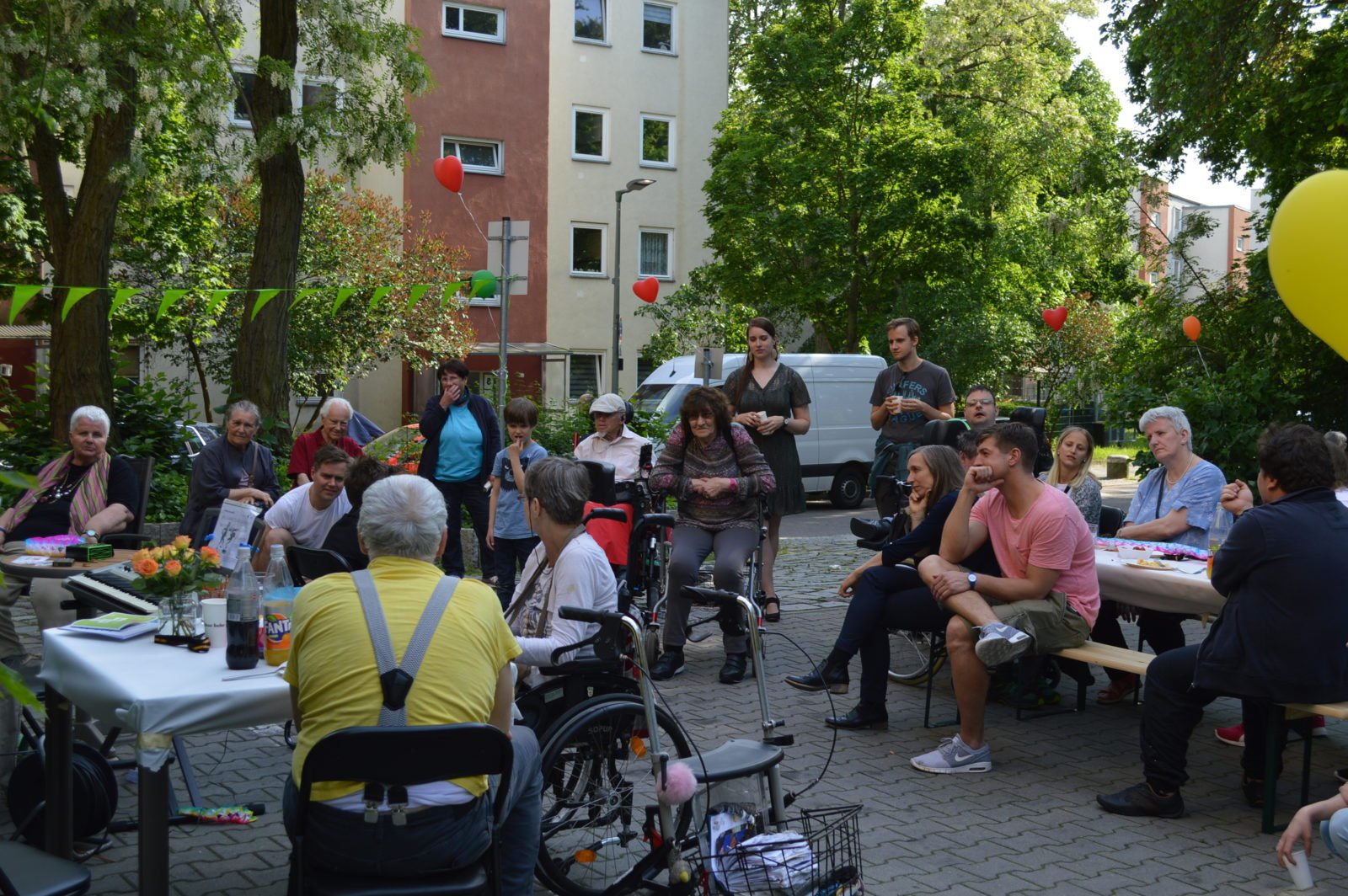 Eine Menschenmenge sitzt und steht an und um Tische vor einer Reihe Wohnhäuser im Hintergrund. Einige Menschen davon im Rollstuhl und andere mit Rollatoren als Gehhilfe. Die Tische sind bunt gedeckt, im Hintergrund sieht man auch überall bunte mit Gas befüllte Luftballons, die zur Dekoration befestigt wurden.