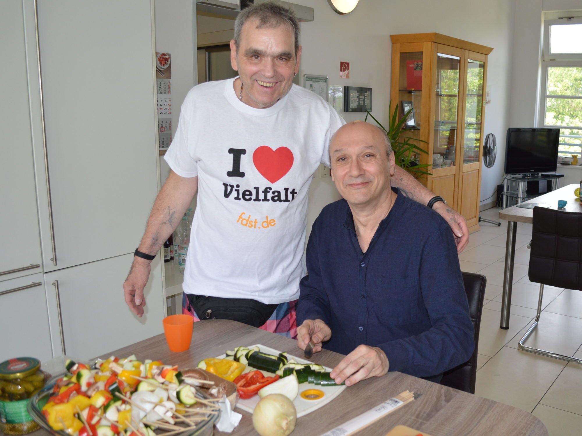 Ein Herr sitzt an einem Tisch und schneidet Gemüse für Grillspieße. Ein anderer Herr steht neben ihm, und trägt ein Shirt mit der Aufschrift I Love Vielfalt. Beide schauen in die Kamera und lächeln.