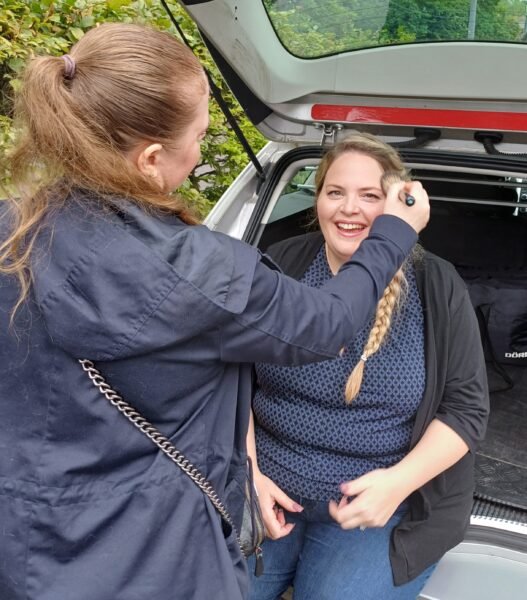 Eine Frau sitzt unter der geöffneten Kofferraum eines silbernen Autos und wird von einer Frau geschminkt