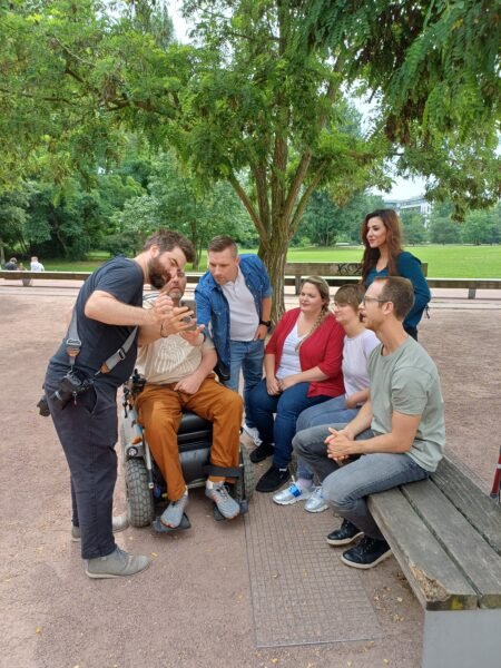 Zwei Frauen und ein Mann sitzen auf einer Parkbank, hinter ihnen steht eine weitere Frau und ein weiterer Mann. Neben ihnen sitzt ein Mann im Rollstuhl und ein Fotograf. Der Fotograf zeigt allen Blder auf seiner Kamera