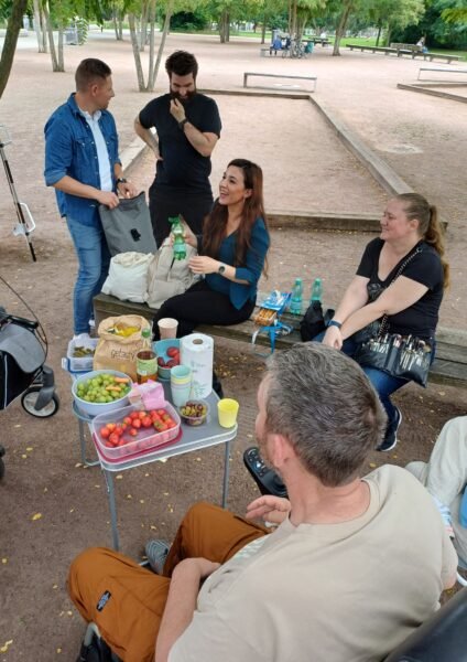 Zwei Frauen sitzen auf einer Parkbank, hinter ihnen stehen 2 Männer. Vor den Frauen steht einer Tisch mit Essen wie Erdbeeren und Weintrauben. Im Vordergrund sitzt ein Mann im Rollstuhl
