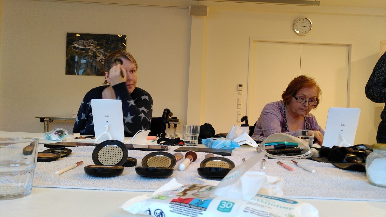 Zwei Frauen, beide im Rollstuhl, sitzen an einem Tisch und tragen Make-up auf.