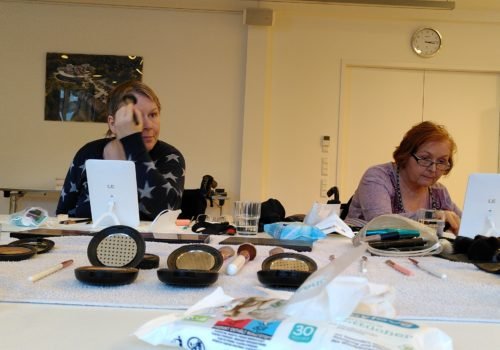 Zwei Frauen, beide im Rollstuhl, sitzen an einem Tisch und tragen Make-up auf.