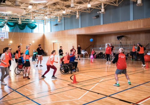 Menschen mit und ohne Behinderung, manche im Rollstuhl, spielen gemeinsam eine Ballsportart in einer Turnhalle.