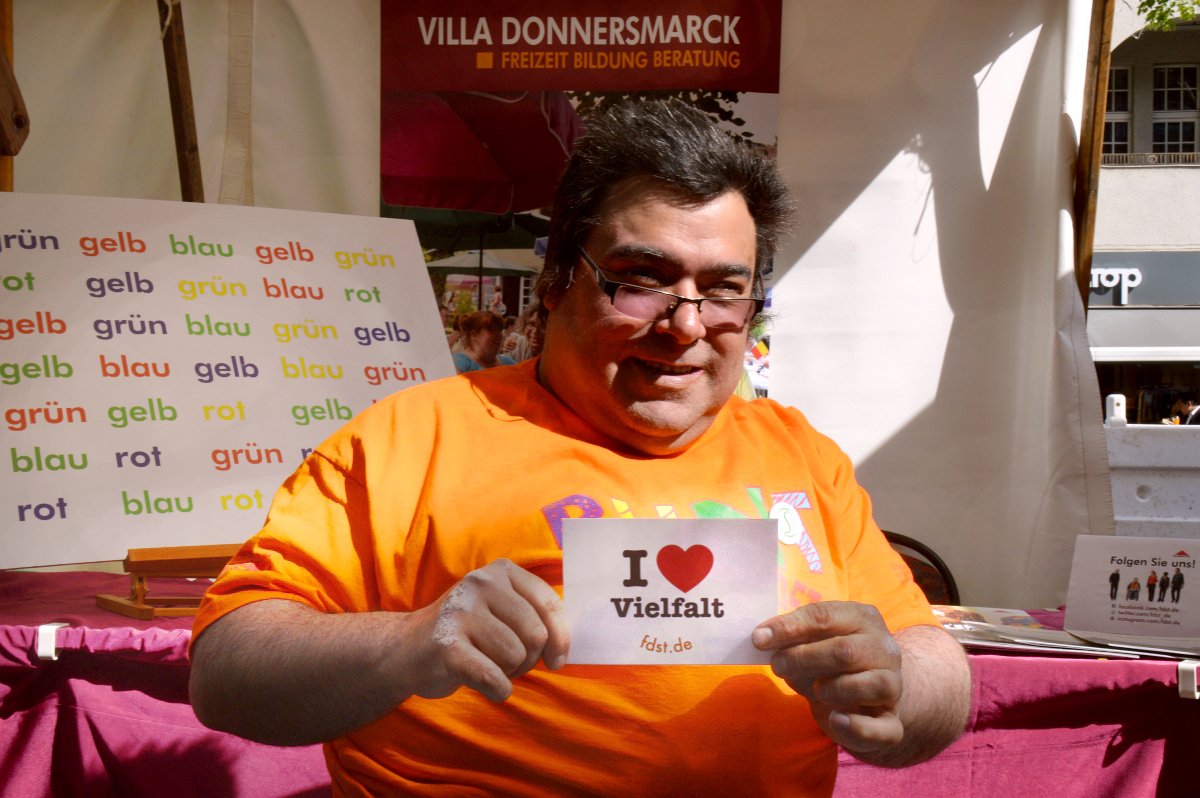 Ein Rollstuhlfahrer mit orangenem T-Shirt zeigt die Karte "I love Vielfalt" in die Kamera