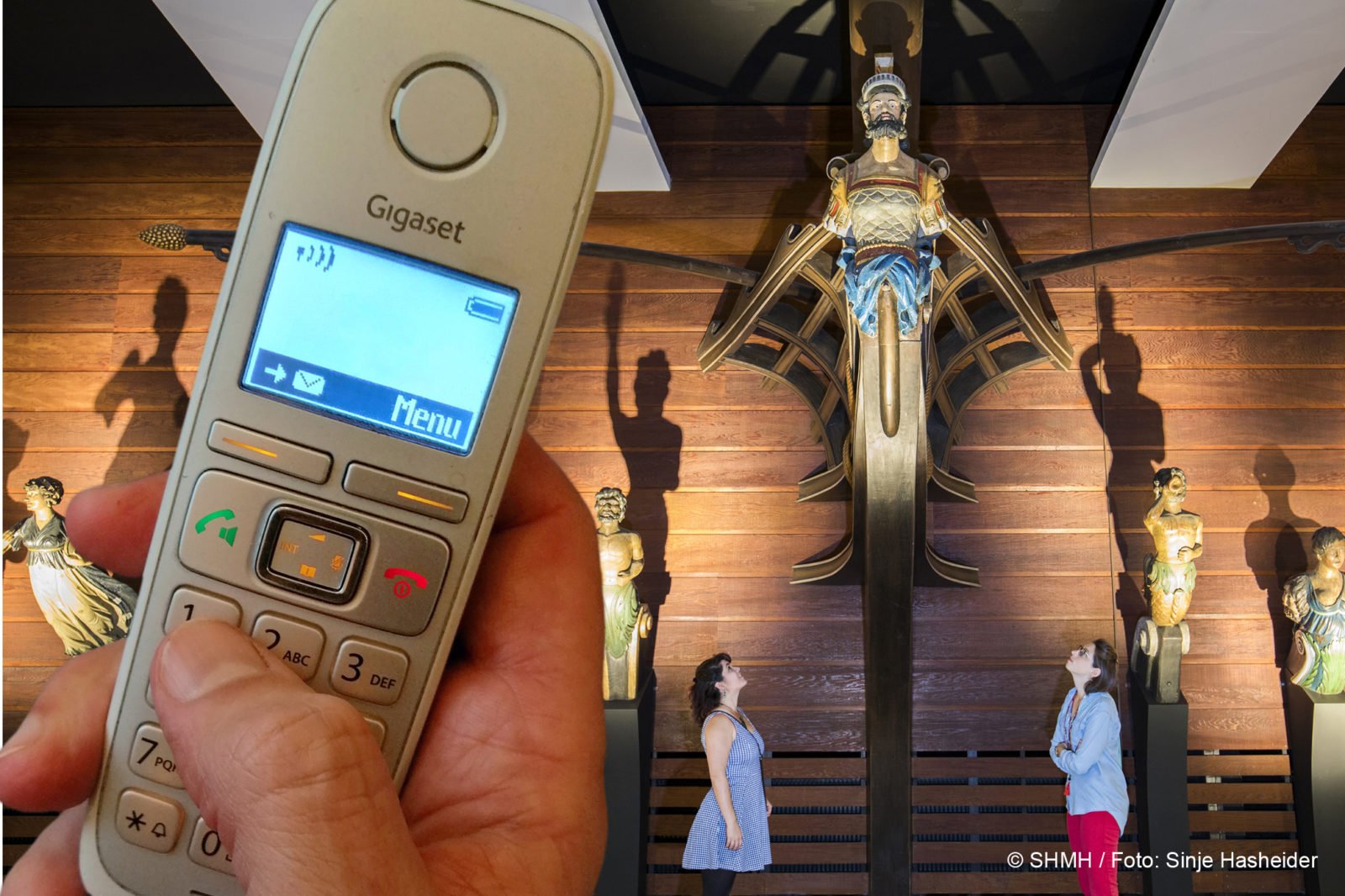 Im Vordergrund eine Hand mit Telefon. Im Hintergrund unterschiedliche Galleonsfiguren historischer Schiffe.