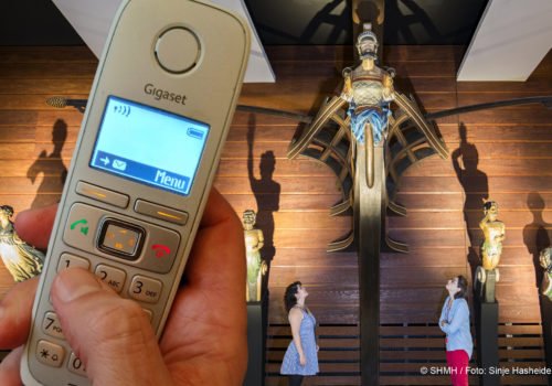 Im Vordergrund eine Hand mit Telefon. Im Hintergrund unterschiedliche Galleonsfiguren historischer Schiffe.