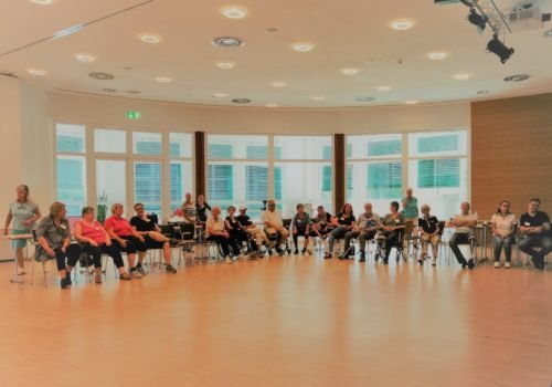 Eine große Gruppe Menschen, teilweise mit Rollstuhl, sitzt in einer großen Halle im Halbkreis. Im Hintergrund große Fenster.