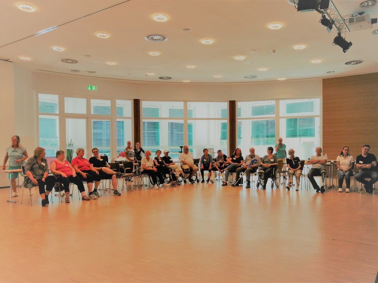 Eine große Gruppe Menschen, teilweise mit Rollstuhl, sitzt in einer großen Halle im Halbkreis. Im Hintergrund große Fenster.