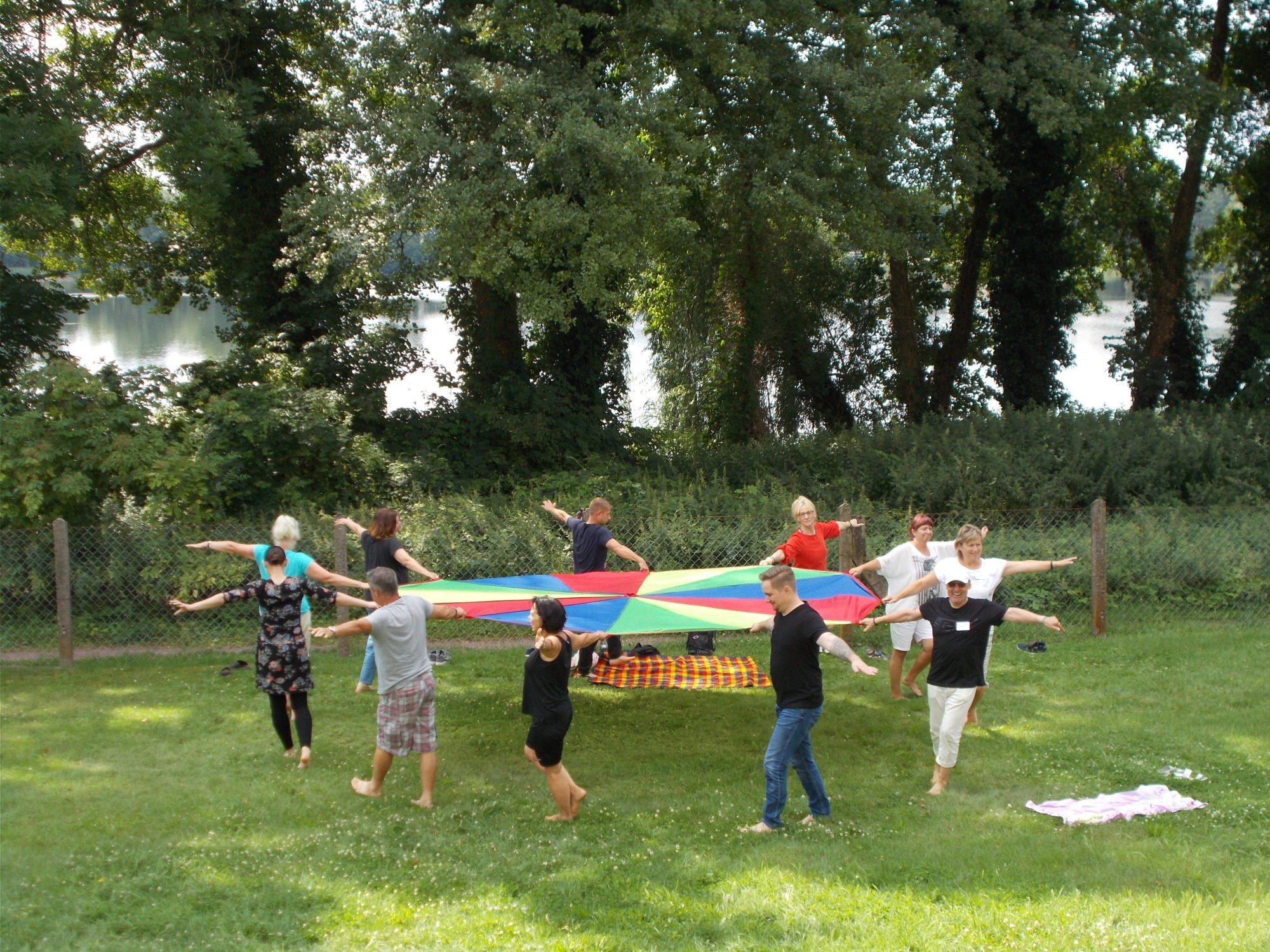 Eine Gruppe Menschen hält auf einer Wiese, jeweils mit dem rechten Arm, gemeinsam ein großes, buntes Tuch und bewegt sich mit dem Tuch im Kreis. Die linken Arme sind nach außen gestreckt. Im Hintergrund sind Bäume und ein See zu sehen.