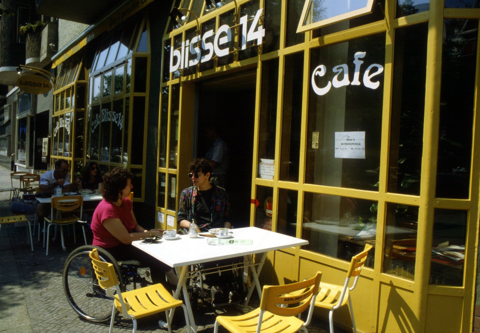 Außenansicht der "blisse 14". Zwei Menschen im Rollstuhl sitzen außen, die Fassade ist gelb. Die Aufschrift "blisse 14" Cafe.