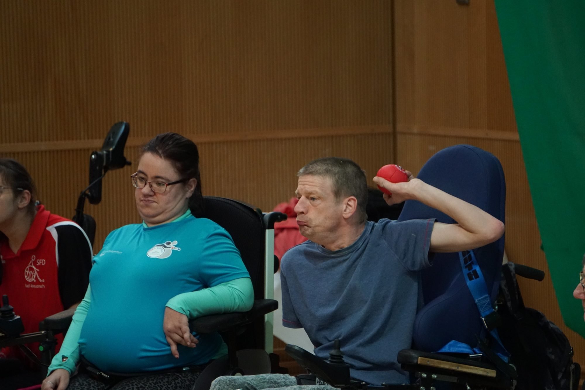 Das Bild zeigt eine Wurfsituation: Ein E-Rollstuhlfahrer ist gerade dabei, einen roten Ball loszulassen. Eine Mitspielerin links neben ihm schaut dabei zu.