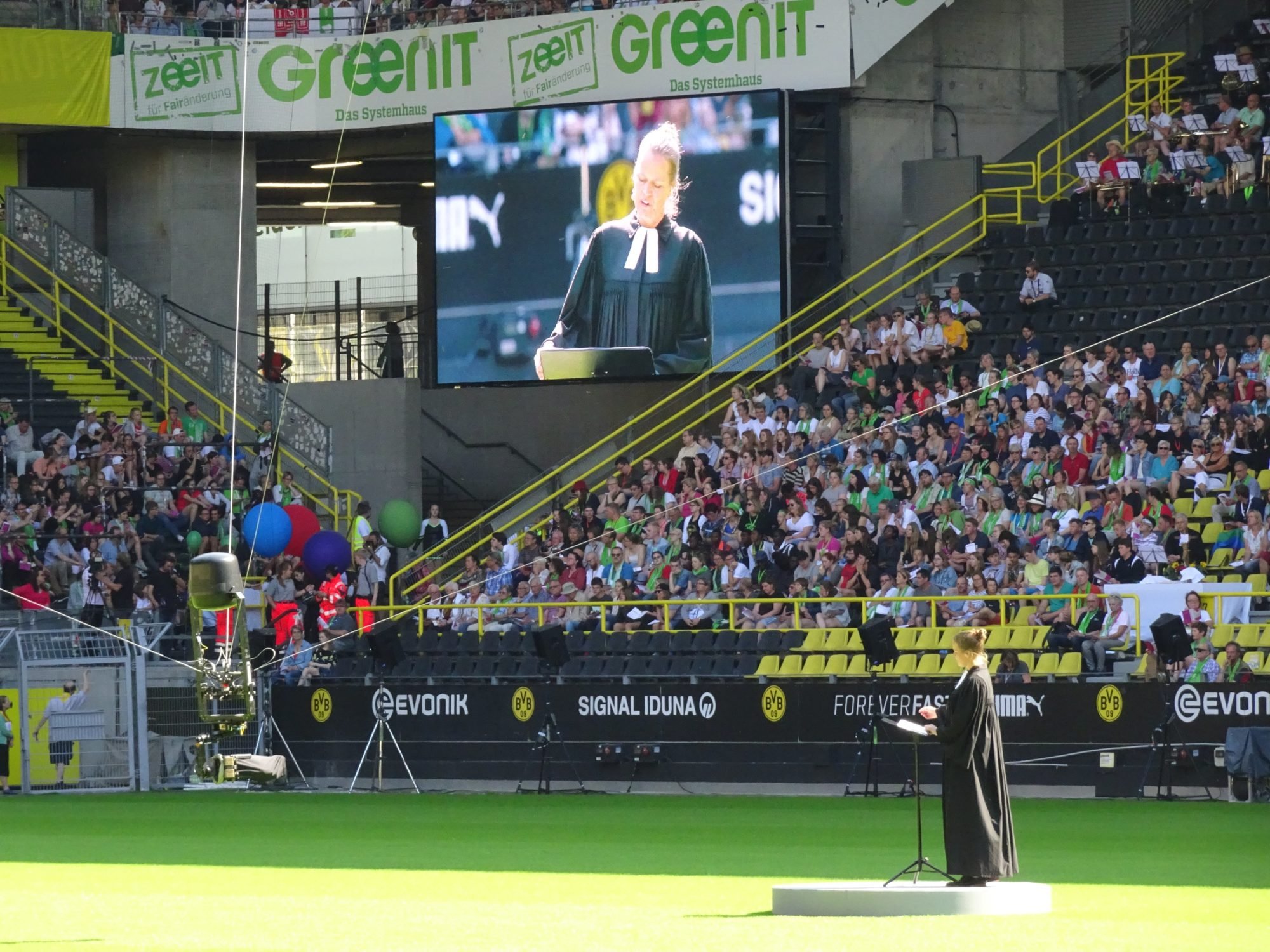 Eine Frau im schwarzen Talar steht auf dem Fußballfeld. Sie hält die Predigt. Im Hintergrund ist Signal Iduna und Evonik zu lesen. Es ist das Stadion von Borussia Dortmund.