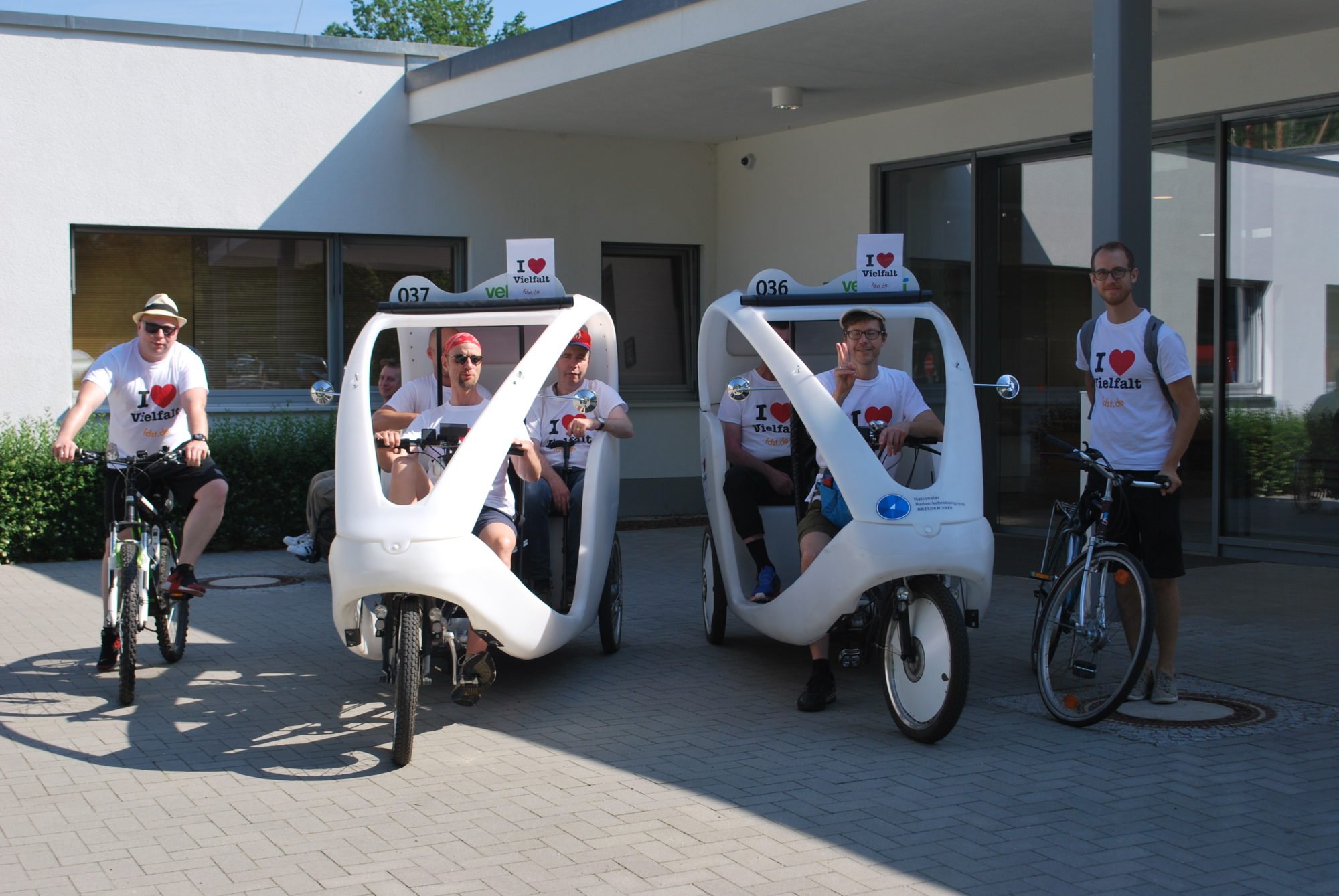 Insgesamt sind sieben Personen zu sehen. Drei fahren mit einer elektrischen Rikscha, zwei Fahren mit einem Fahrrad. Sie stehen vor dem P.A.N. Zentrum und tragen ein "I love Vielfalt" T-Shirt.