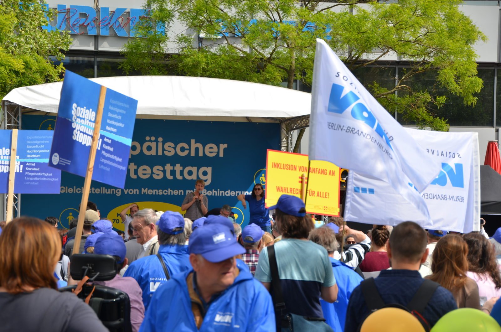 Protesttag: Blick auf die Kundgebungsbühne des Europäischen Protesttags zur Gleichstellung von Menschen mit Behinderung. Im Vordergrund sind jede Menge Demonstranten, Transparente und Schilder im Bild zu sehen.