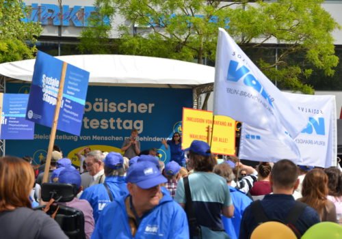 Protesttag: Blick auf die Kundgebungsbühne des Europäischen Protesttags zur Gleichstellung von Menschen mit Behinderung. Im Vordergrund sind jede Menge Demonstranten, Transparente und Schilder im Bild zu sehen.