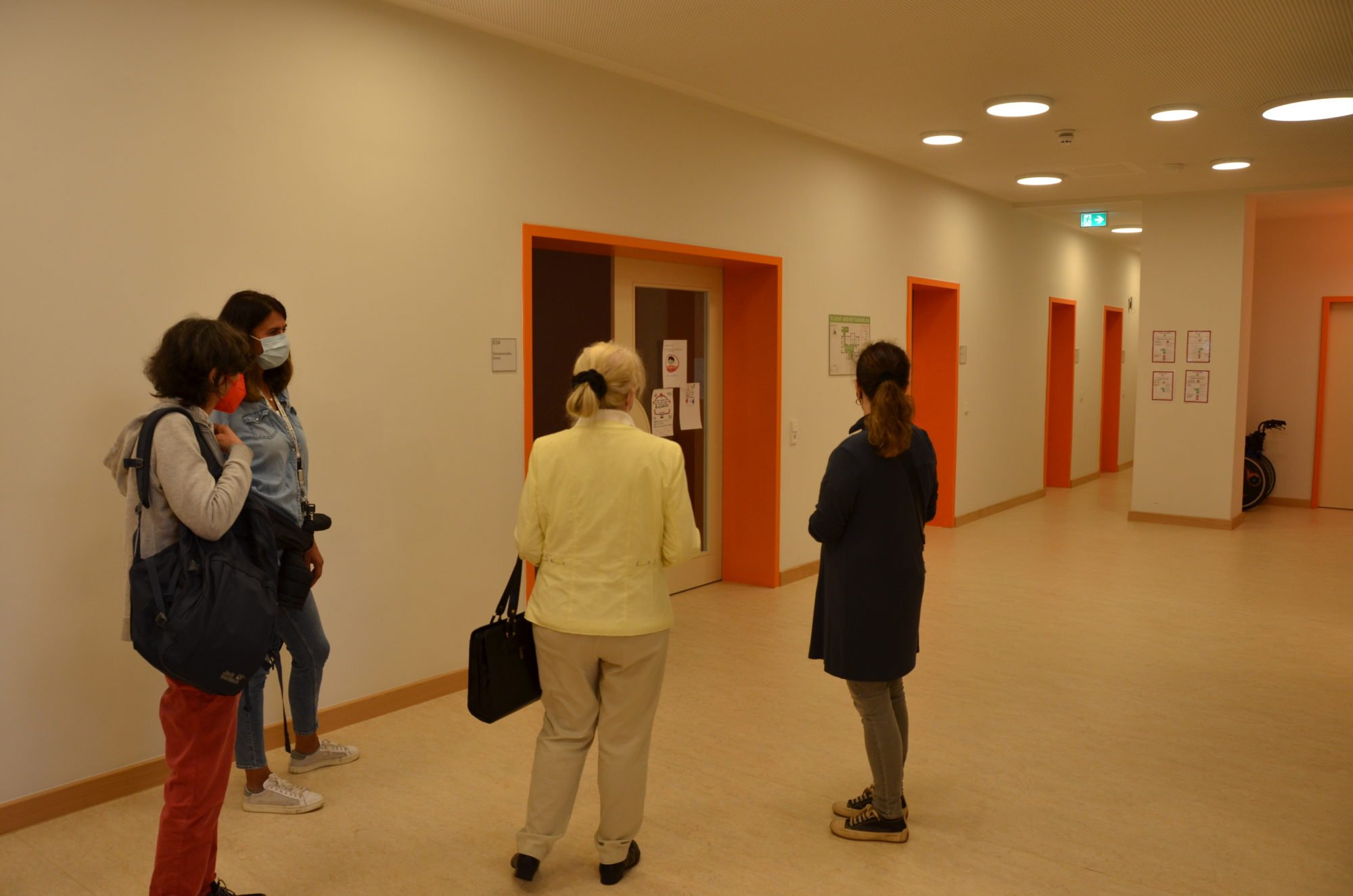 Vier Frauen bei der Begehung des Frauenhaus. Deutlich sichtbar sind orangene Elemente im im Flur, die die Etage farblich kennzeichnen.