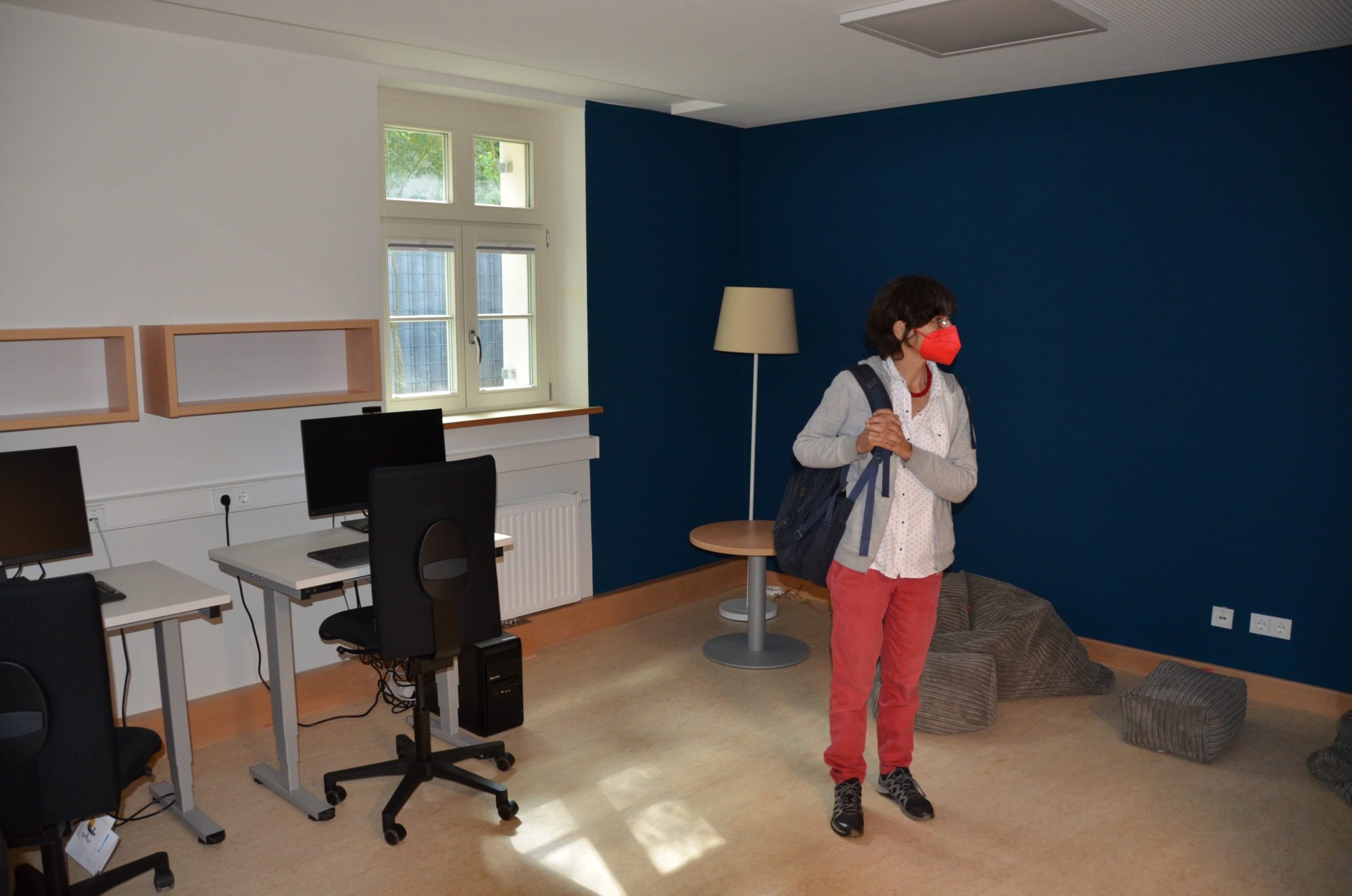 Eine Frau betrachtet den Raum. Links: Computertische mit Stühlen, PCs und Monitoren. Rechts: eine Chillout-Ecke mit Sitzkissen und Lampe.