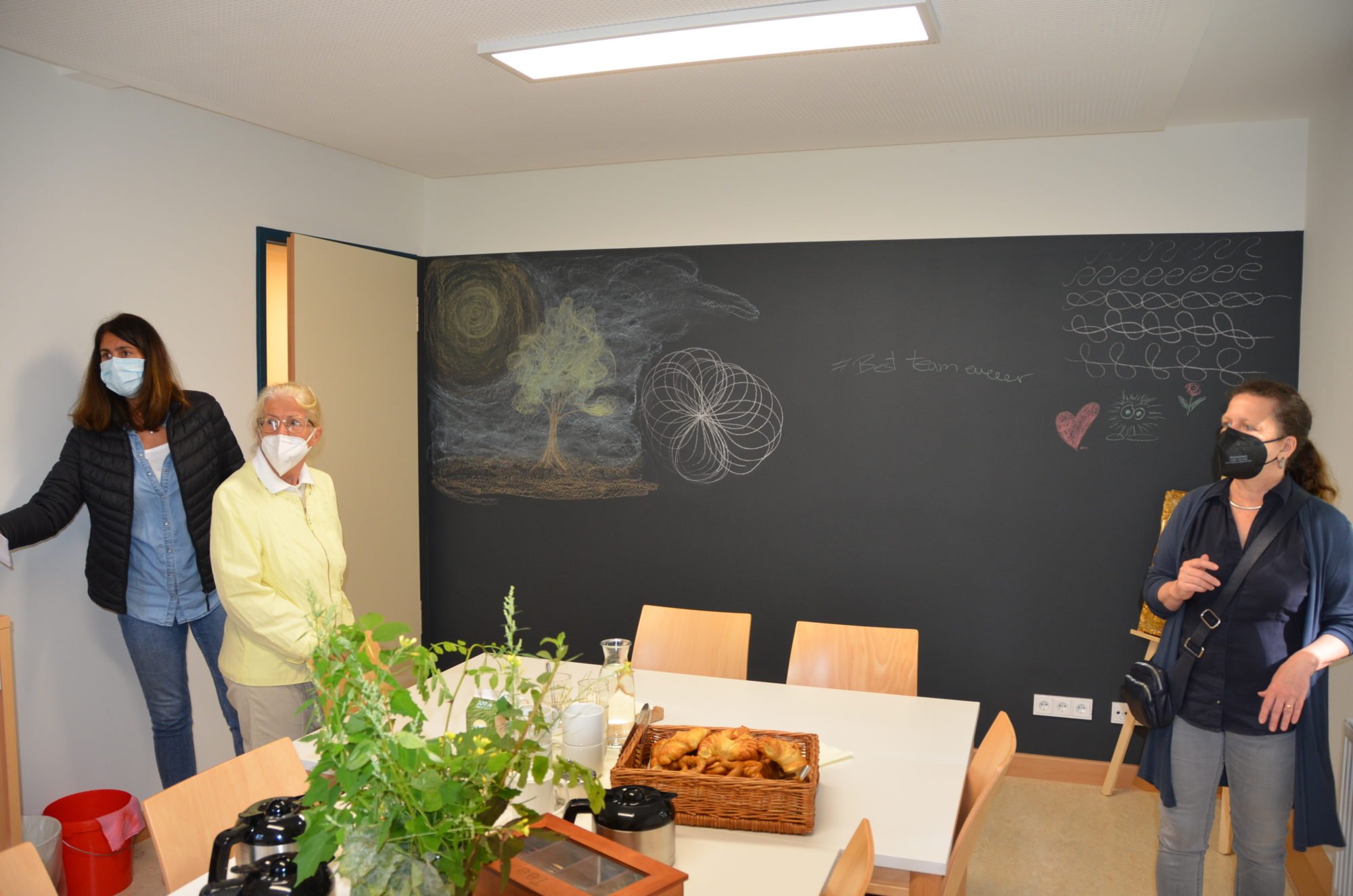 Ein freundlich eingerichteter Raum. Im Hintergrund eine bemalte Schiefertafelwand. Auf dem Tisch in der Raummitte stehen Pflanzen und ein Korb mit Gebäckstücken.