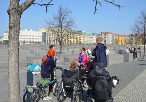 Eine Gruppe Menschen, darunter drei Rollstuhlfahrerinnen und -fahrer sowie eine Frau mit Fahrrad, steht gemeinsam vor dem Holocaust-Mahnmal in Berlin-Mitte. Der Himmel ist klar und die Sonne scheint.