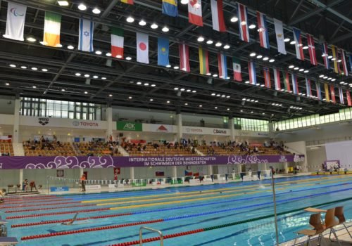 Blick in die Schwimmhalle: Vorne das Becken in dem gerade ein Wettkampf stattfindet, im Hintergrund die Ränge mit Zuschauern und darüber die Flaggen aller teilnehmenden Nationen.
