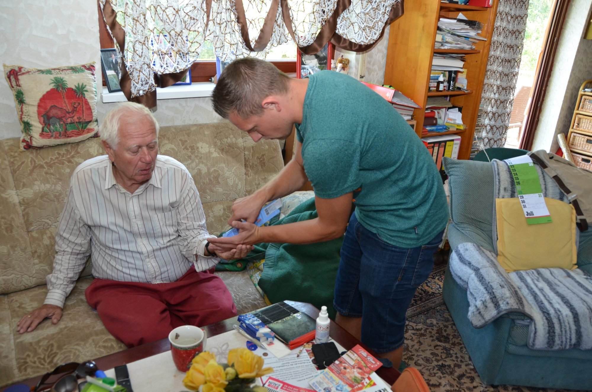 Marco, rechts im Bild ist leicht nach vorne gebeugt und gibt einem älteren Herren, der auf seinem Sofa sitzt, seine Medikamente in die Hand.