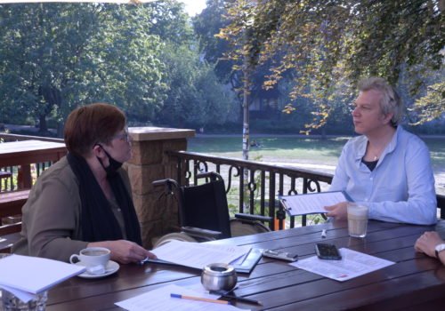 Sabine Haller und Martin Küster in der Interviewsituation im Freien an einem Holztisch.