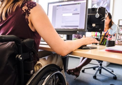 Eine junge Frau im Rollstuhl sitzt an ihrem Bildschirmarbeitsplatz. Im Hintergrund ist eine Kollegin zu sehen, die ebenfalls gerade an ihrem Bildschirm arbeitet.
