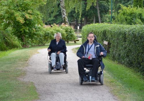 Ein E-Rolli-Fahrer und eine E-Rolli-Fahrerin fahren auf einem kleinen Fahrradweg auf die Kamera zu und lächeln.