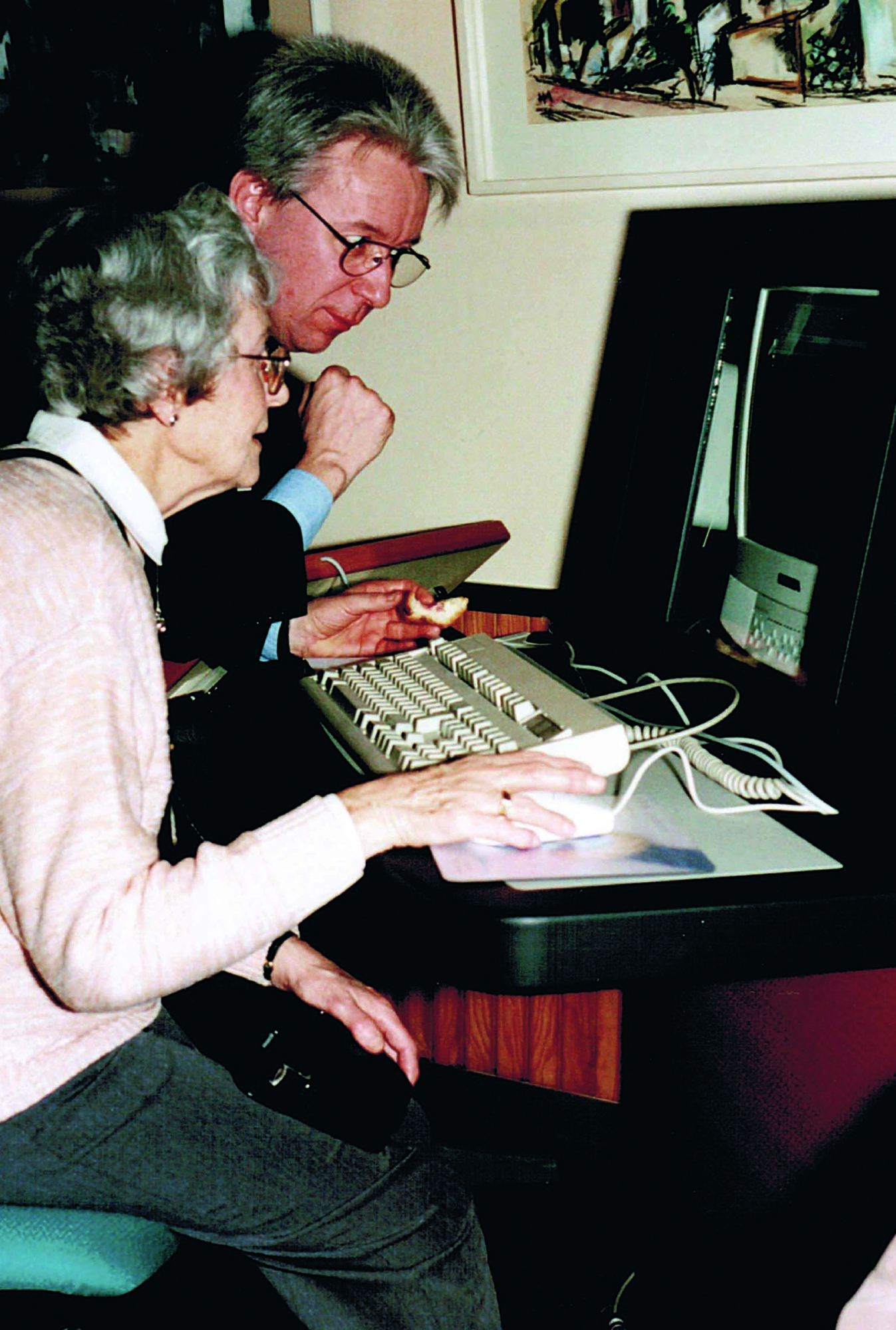 Zu sehen ist ein Mann, der sich über die Schulter einer älteren Dame lehnt. Die Dame schaut auf einen alten PC und hat die rechte Hand an der Maus.