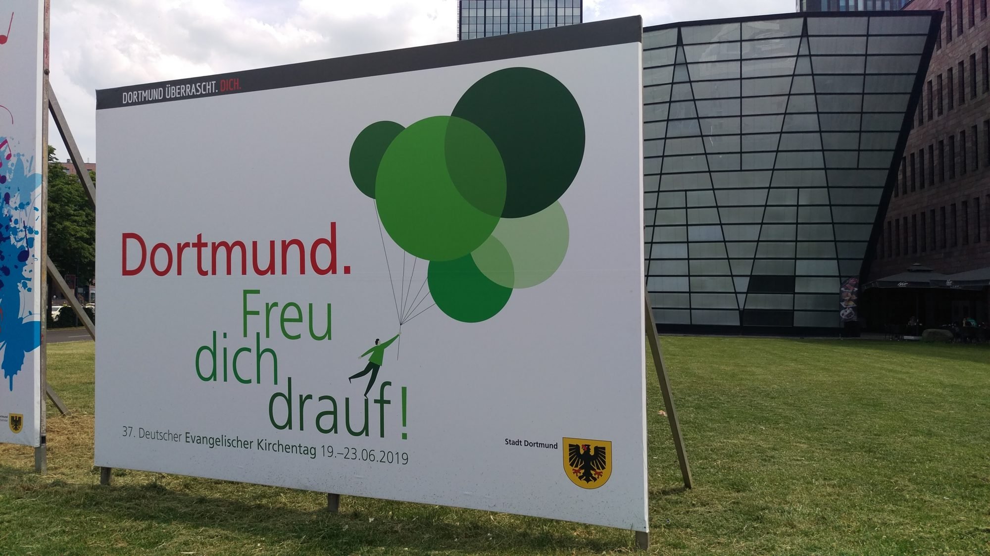 Ein Werbeplakat für den 37. Deutschen Evangelischen Kirchentag mit der Aufschrift "Dortmund. Freu dich drauf!" mit einer gezeichneten Figur, die an Luftballons hängt und davonfliegt sowie einem Wappen der Stadt Dortmund im unteren rechten Eck.