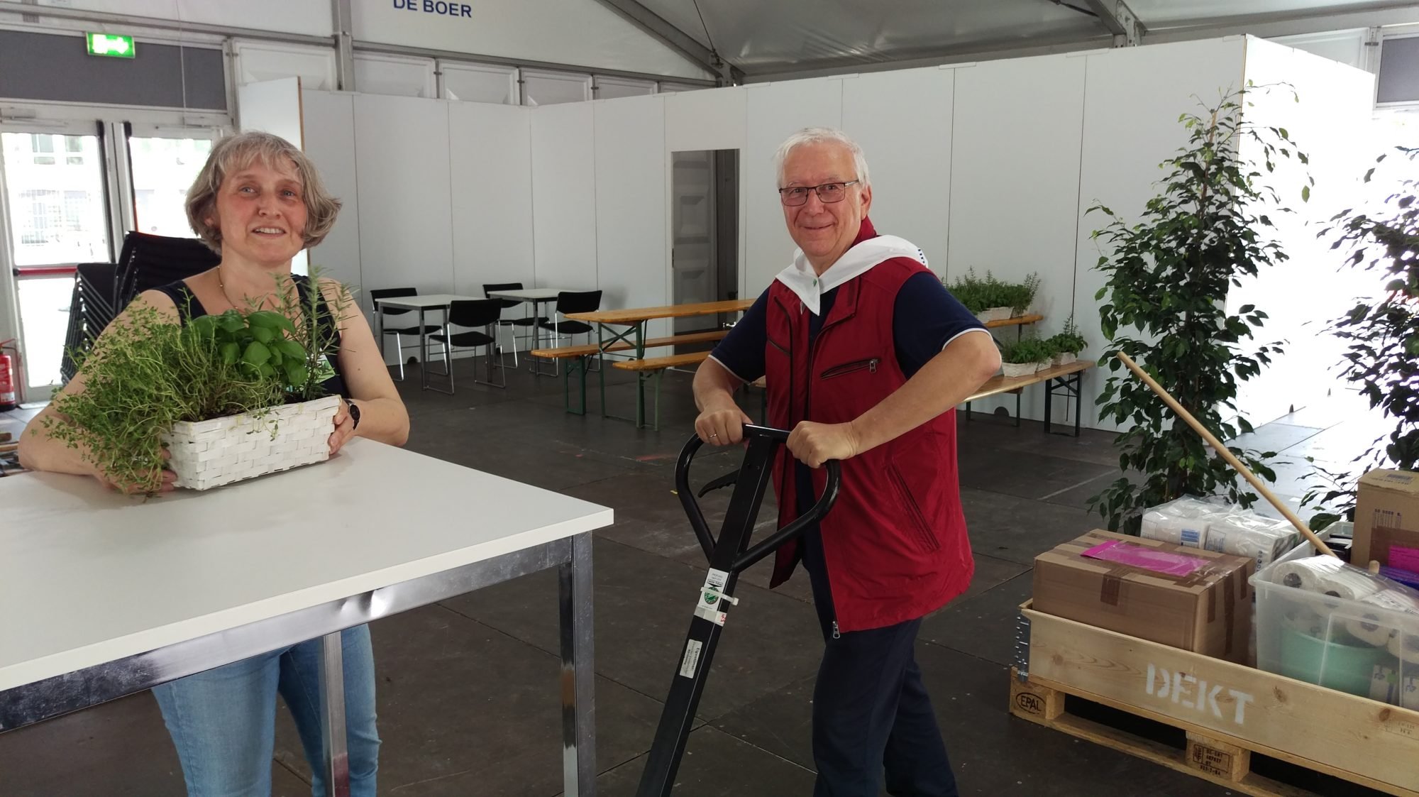 Links steht eine Frau an einem Tisch und stellt eine Pflanze ab. Rechts von ihr steht Thomas Golka mit den Händen an einem Hubwagen, mit dem er Einrichtungsgegenstände transportiert. Beide schauen lächelnd Richtung Kamera.