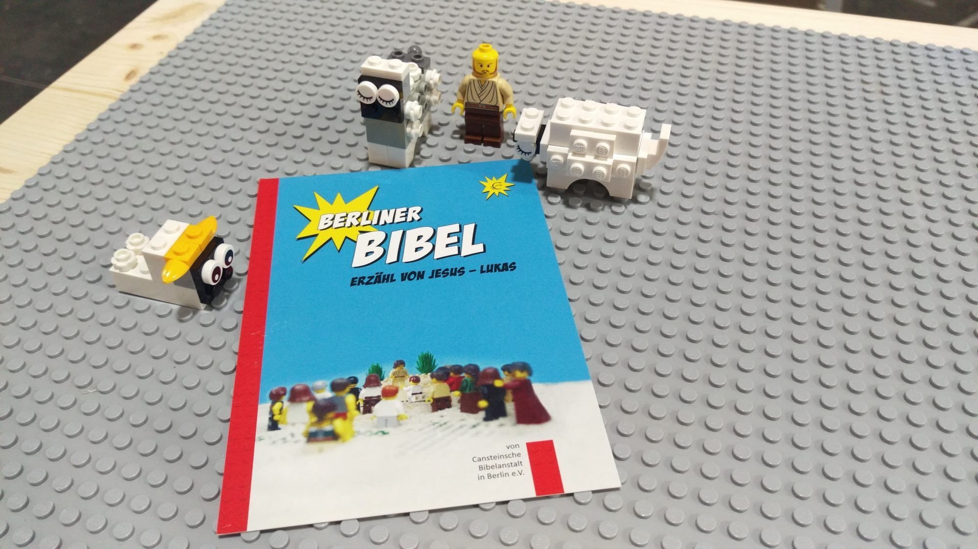 Eine Lego-Miniatur und drei Schafe aus Legosteinen auf einem grauen Lego-Untergrund. Darauf liegt ein Flyer mit der Aufschrift: "Berliner Bibel. Erzähl von Jesus - Lukas"