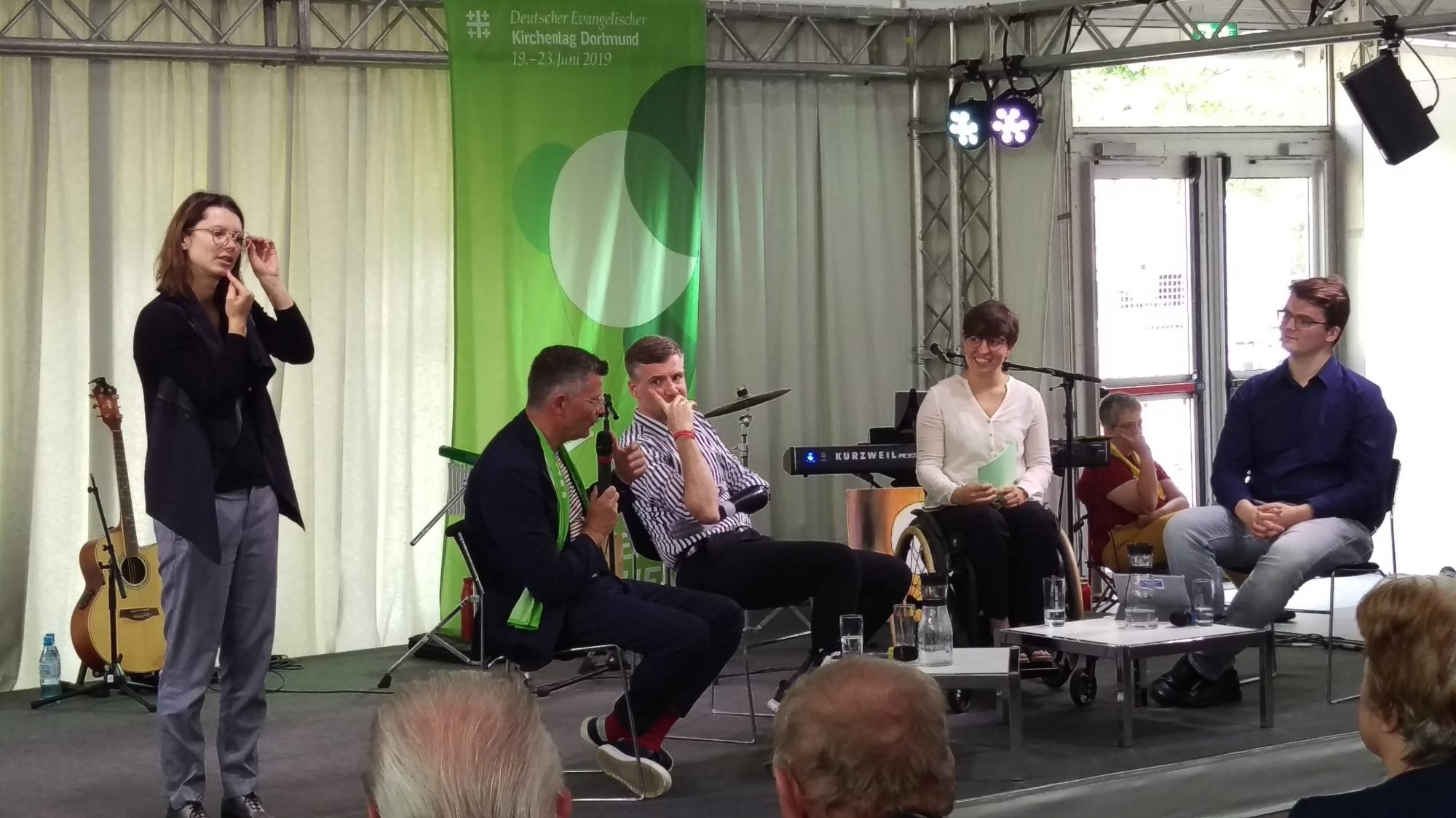 Ein Gruppe sitzt auf dem Podium und diskutiert. Links ein älterer Herr, neben ihm ein junger Mann, daneben eine junge Frau im Rollstugl und ganz rechts ein junger Mann mit Brille. Links im Bild ist eine Gebärdensprach-Dolmetscherin zu sehen.