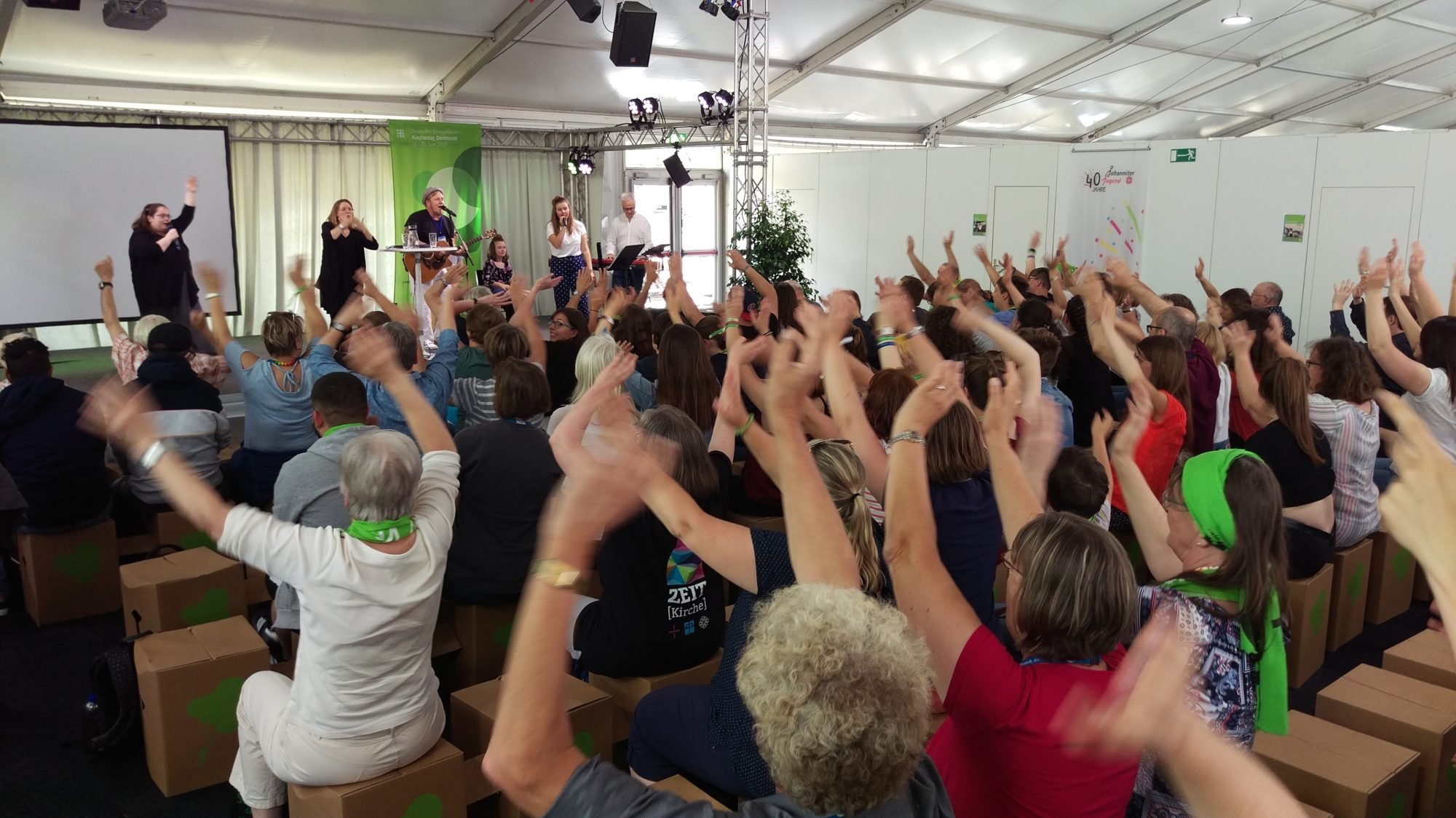 Foto über die Köpfe des Publikums hinweg. Alle Hände gehen nach oben. Auf der Bühne spielt die Band.