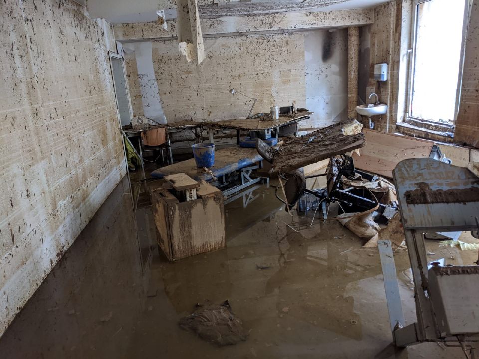 Ein komplett von der Flut zerstörter Raum. Die Tapete ist komplett von den Wänden verschwunden, im Raum verteilt liegt zerstörtes Mobiliar und dreckiges Wasser steht noch etwa Knöcheltief im Raum.